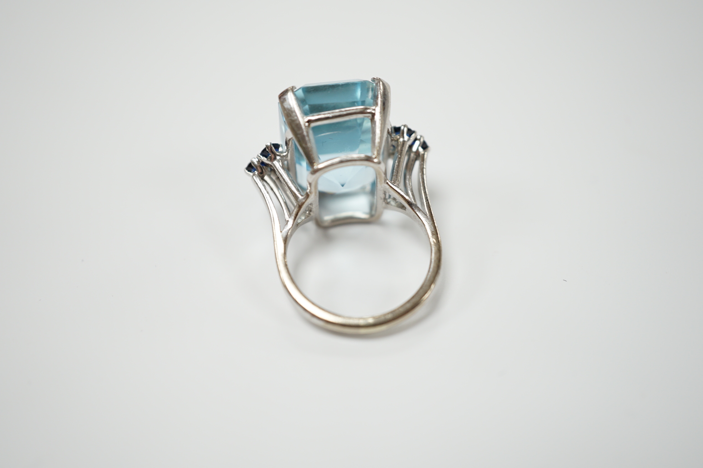An 18ct white metal and single stone emerald cut aquamarine set dress ring, with six stone sapphire set shoulders, size L, gross weight 11.8 grams.